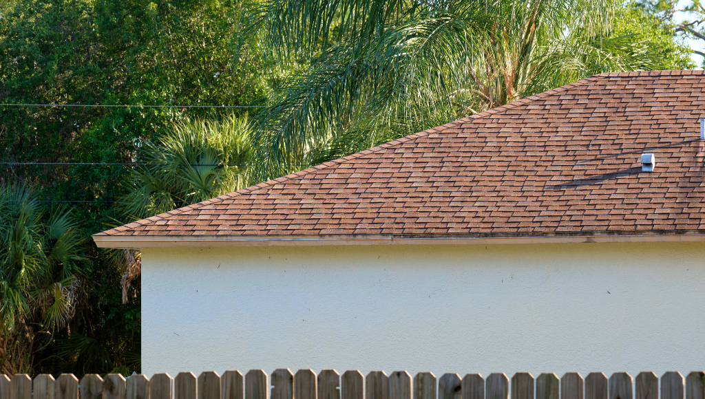 Close-up of a section of hip roof, covered in reddish-brown asphalt shingles. A wooden picket fence sits in the foreground, and foliage, including palm trees, in the background.