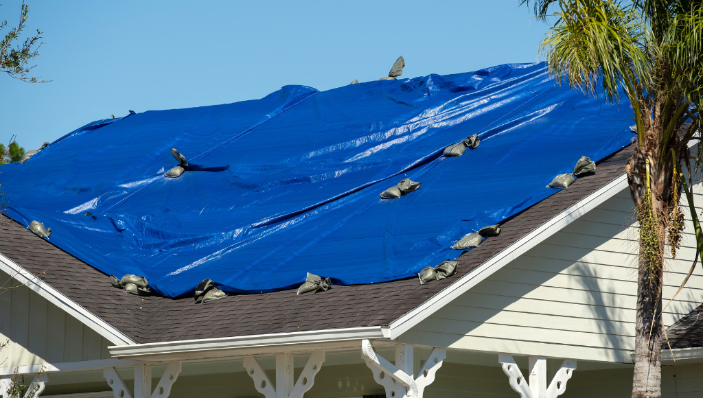 A bright blue tarp is held down by sandbags, temporarily covering a brown shingled roof.